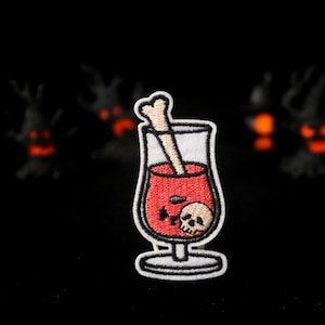May include: Embroidered patch featuring a cocktail in a glass with a bone stirrer and a skull. The drink is red, and the patch has a white border. The background is black with blurred Halloween decorations.