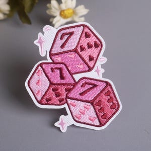 May include: Embroidered patch featuring three pink dice with the number 7 on each face. The dice are decorated with heart shapes and surrounded by star accents. The patch is on a white background.