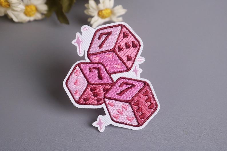 K&ouml;nnte beinhalten: Gestickter Aufn&auml;her mit drei rosa W&uuml;rfeln, die jeweils die Zahl 7 zeigen. Die W&uuml;rfel sind mit Herzmotiven und Sternen verziert. Der Aufn&auml;her befindet sich auf einem wei&szlig;en Hintergrund.