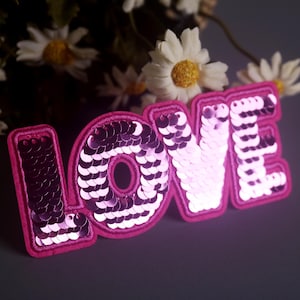 May include: Pink fabric patch with the word "LOVE" spelled out in large, glittery letters. Each letter is covered in pink sequins.