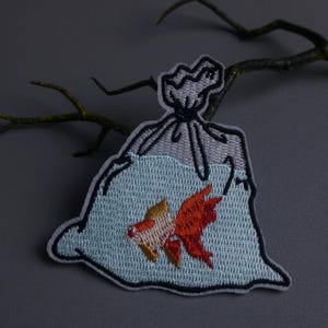 May include: Embroidered patch featuring a goldfish in a plastic bag. The patch has a light blue background with a black outline. The goldfish is orange and yellow. The bag is gray and tied at the top.