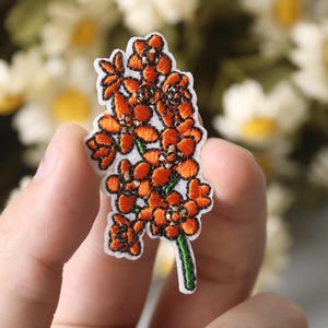 Puede incluir: Parche bordado de flores naranja con tallo y hojas verdes. El parche está sobre un fondo blanco.