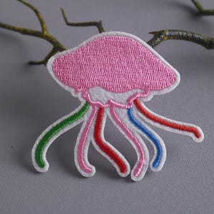 May include: Pink embroidered jellyfish patch with white outline and rainbow colored tentacles.