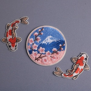 May include: Embroidered patches featuring Japanese motifs. Includes two koi fish patches in red, white, and brown, and a circular patch with Mount Fuji, cherry blossoms, and a blue sky background.
