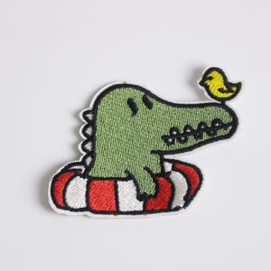 May include: Embroidered patch featuring a green alligator wearing a red and white striped inner tube. A yellow bird sits on the alligator's head. The patch has a white border and is likely for clothing or accessories.