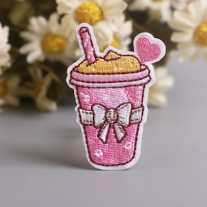 Pode incluir: Remendo bordado de uma bebida rosa com canudo e coração. A bebida tem a parte superior amarela e um laço branco. O remendo está em uma superfície cinza com flores brancas ao fundo.