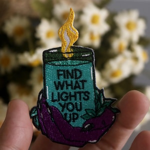 May include: Embroidered patch featuring a teal candle with a yellow flame, held by a purple hand. The text "FIND WHAT LIGHTS YOU UP" is embroidered on the candle. The patch is set against a blurred background of white flowers.