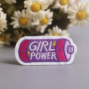 May include: Embroidered patch with the words "GIRL POWER" in white and purple, designed to resemble a battery. The patch has a white border and is set against a gray background with daisies.