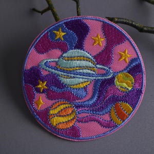 May include: A pink, purple, and blue embroidered patch with a swirling design featuring planets and stars. The patch is circular and has a white background.