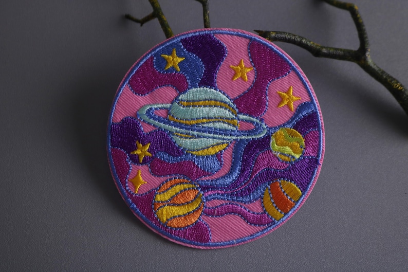May include: A pink, purple, and blue embroidered patch with a swirling design featuring planets and stars. The patch is circular and has a white background.