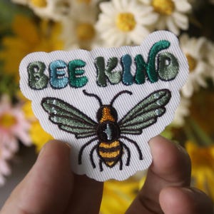 May include: A white embroidered patch with a bee and the text "Bee Kind" in green and blue lettering.