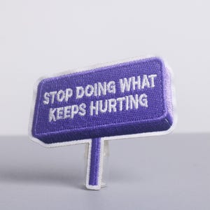 Puede incluir: Un parche bordado morado en forma de letrero con el texto "STOP DOING WHAT KEEPS HURTING" en blanco. El parche tiene un borde blanco y un tallo blanco.
