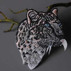 May include: Embroidered patch of a snow leopard with blue eyes. The patch is detailed with black and white fur and a pink nose.