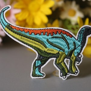 May include: A colorful embroidered iron-on patch featuring a dinosaur with a blue, green, yellow, and orange design.