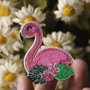 Könnte beinhalten: Rosa gestickter Flamingo-Aufnäher mit grünen Blättern und rosa Blüten.