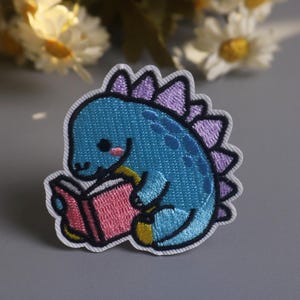 May include: Embroidered patch featuring a blue dinosaur reading a pink book. The dinosaur has purple spines and is outlined in black. The patch is on a gray background with flowers in the background.