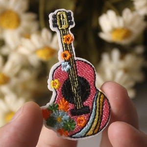 May include: A pink embroidered guitar patch with floral details. The guitar has a black and white striped design on the body and a black and white striped design on the neck. The patch is surrounded by white daisies.