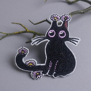 May include: Embroidered black cat patch with a daisy crown and daisies on its tail. The cat has purple eyes and whiskers. The patch is on a gray background with a branch.