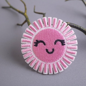 May include: A pink embroidered sun with a smiling face and eyelashes. The sun has white rays.