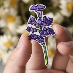 Puede incluir: Parche bordado de flores moradas con tallo verde. El parche se sostiene en una mano sobre un fondo borroso de flores blancas.