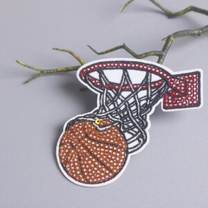Peut inclure: &Eacute;cusson brod&eacute; repr&eacute;sentant un ballon de basket passant &agrave; travers un panier. L'&eacute;cusson pr&eacute;sente un ballon de basket marron &agrave; pois blancs, un panier rouge et blanc et un filet noir. L'&eacute;cusson est sur fond gris avec une branche.