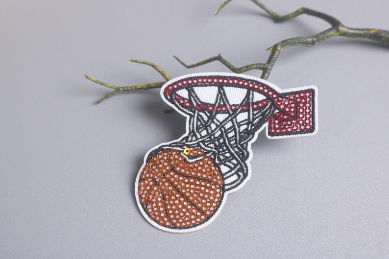 Peut inclure: &Eacute;cusson brod&eacute; repr&eacute;sentant un ballon de basket passant &agrave; travers un panier. L'&eacute;cusson pr&eacute;sente un ballon de basket marron &agrave; pois blancs, un panier rouge et blanc et un filet noir. L'&eacute;cusson est sur fond gris avec une branche.