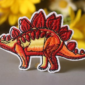 May include: Embroidered iron-on patch of a red and yellow dinosaur with a spiky back.