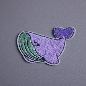 Può includere: Toppa ricamata raffigurante una balena dei cartoni animati nei toni del viola e del verde. La balena ha una coda a forma di cuore e un viso sorridente. La toppa è bordata di bianco su sfondo grigio.