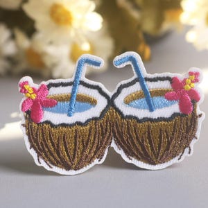 May include: Embroidered patch featuring two coconut drinks with blue straws, white coconut flesh, and brown husks. Each coconut has a pink flower with yellow accents. The patch is designed for clothing or accessories.