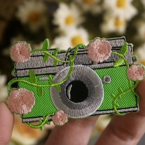 May include: Green embroidered camera patch with pink flowers and vines. The camera has a silver lens and a black aperture.