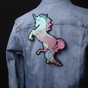 Puede incluir: Una chaqueta vaquera con un parche de unicornio iridiscente rosa y azul cosido en la espalda.