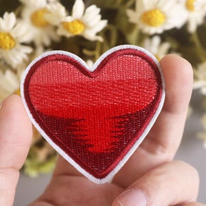 May include: A red heart-shaped embroidered patch with a white outline. The patch has a gradient of red shades, with a darker red at the center and a lighter red at the edges.