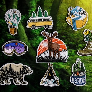 May include: A collection of embroidered patches with outdoor and travel themes. Designs include a lightbulb with mountains, a yellow van, a mountain scene in a mug, ski goggles, a moose, a boot with flowers, a bear, a tent, and a mug with the word "EXPLORE".