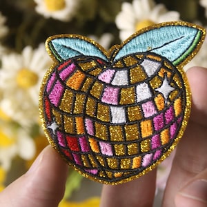 May include: A gold, pink, orange, and white embroidered iron-on patch shaped like a peach with a disco ball design.