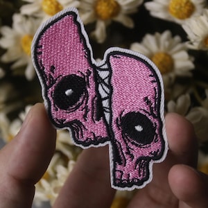Könnte beinhalten: Rosa gestickter Patch mit zwei Schädeln mit schwarzen Augen und Details, die mit einem weißen Faden zusammengenäht sind.