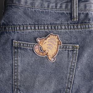 Könnte beinhalten: Ein dunkelgrauer Jeansstoff mit einer Gesäßtasche, auf der ein gestickter Aufnäher einer braunen getigerten Katze von hinten zu sehen ist. Die Katze ist mit einem geschwungenen Schwanz und sichtbaren Streifen mittig auf der Tasche platziert.