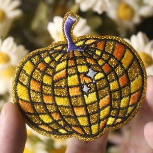 Può includere: Un toppa da stirare a forma di zucca di colore oro e arancione con contorno nero. La toppa ha un motivo a scacchi e due stelle bianche.