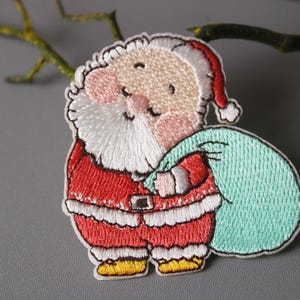 以下が含まれることがあります： 白いひげと赤いスーツを着た漫画のサンタクロースが、水色の袋を持っている刺繍パッチ。サンタクロースは赤い帽子をかぶって笑顔です。パッチは灰色の背景にあります。