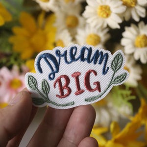 以下が含まれることがあります： 緑の葉と「Dream Big」という文字が青と赤で刺繍された白いパッチ