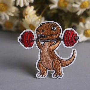 May include: Embroidered patch featuring a cartoon Tyrannosaurus Rex lifting a barbell. The dinosaur is brown with a white outline, and the barbell weights are red. The patch is designed for application on clothing or accessories.