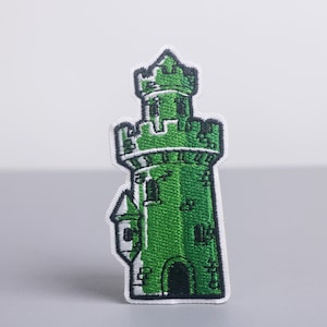 May include: Embroidered patch featuring a green castle tower with a white border. The patch has a detailed design with windows and crenellations. The castle is a vibrant green color, and the patch is likely for clothing or accessories.