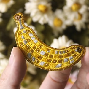 May include: A yellow and white embroidered banana patch with a glittery finish. The patch is ideal for adding a fun and quirky touch to clothing, bags, or other accessories.
