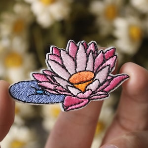 May include: A pink and white embroidered flower patch with a blue leaf. The flower has a yellow center and is surrounded by pink petals.
