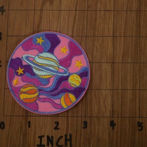 May include: A pink, purple, and yellow embroidered patch with a space theme. It features a planet with rings, stars, and swirling clouds.