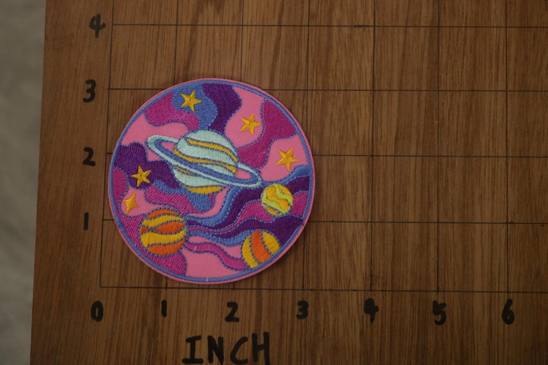 May include: A pink, purple, and yellow embroidered patch with a space theme. It features a planet with rings, stars, and swirling clouds.