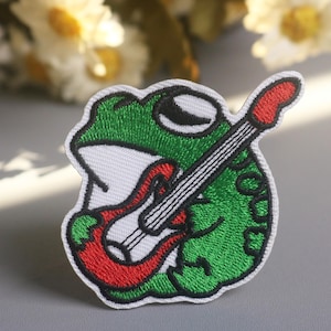 May include: Embroidered patch featuring a green frog playing a red guitar. The frog has a white belly and black eyes. The patch is outlined in black and has a white border. A fun accessory for clothing or bags.