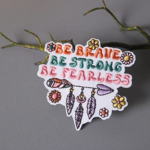 Peut inclure: Patch brodé blanc avec le texte "Be Brave Be Strong Be Fearless" en lettres colorées. Le patch présente une flèche stylisée avec des plumes et des accents floraux.