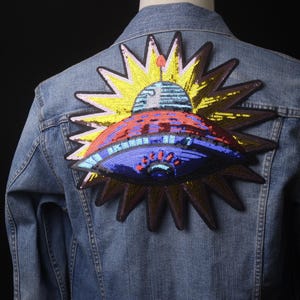 Puede incluir: Una chaqueta vaquera con un parche de lentejuelas de colores de un OVNI volando sobre un sol con rayos. El parche es una mezcla vibrante de azul, rojo, amarillo y dorado.