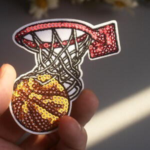 Peut inclure: &Eacute;cusson de panier de basket-ball avec un ballon. Le cerceau est rouge avec un bord blanc et le filet est noir. Le ballon est marron et dor&eacute;, avec une bordure blanche. L'&eacute;cusson est fait de paillettes.