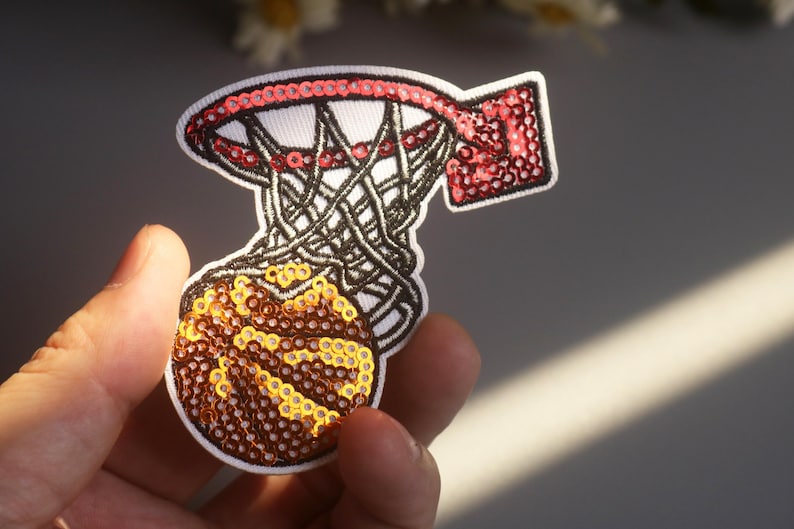 Peut inclure: &Eacute;cusson de panier de basket-ball avec un ballon. Le cerceau est rouge avec un bord blanc et le filet est noir. Le ballon est marron et dor&eacute;, avec une bordure blanche. L'&eacute;cusson est fait de paillettes.